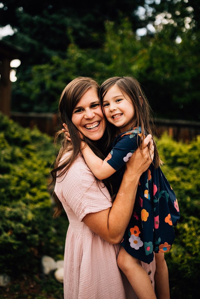 A mother holds her daughter up in their backyard as they both smile at the camera