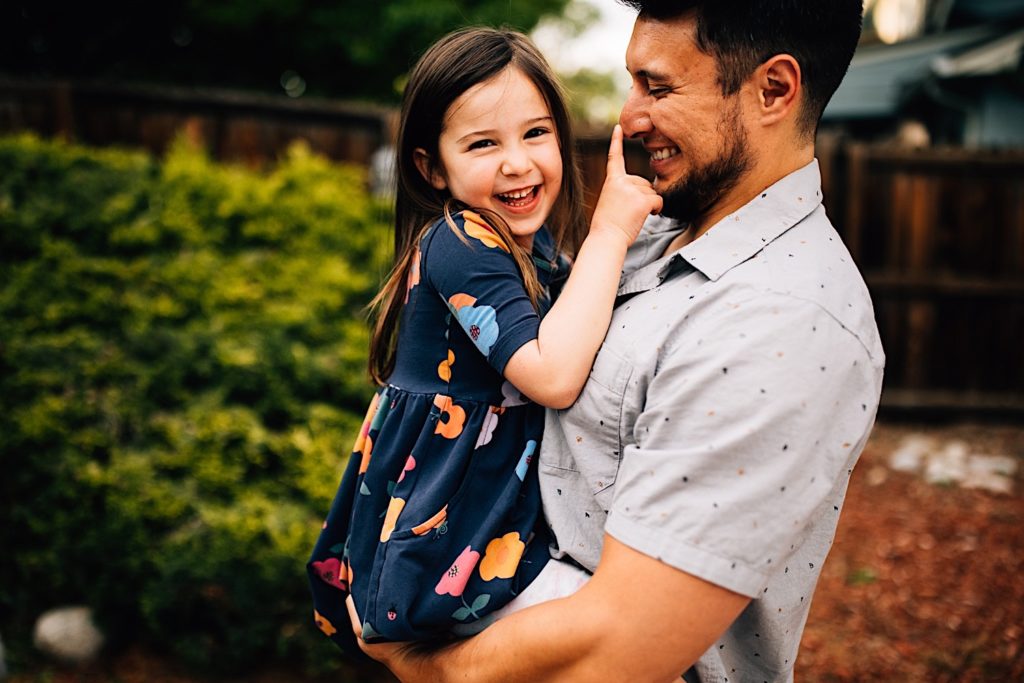 A father holds his daughter, they're both smiling as the daughter pokes her fathers nose and laughs at the camera