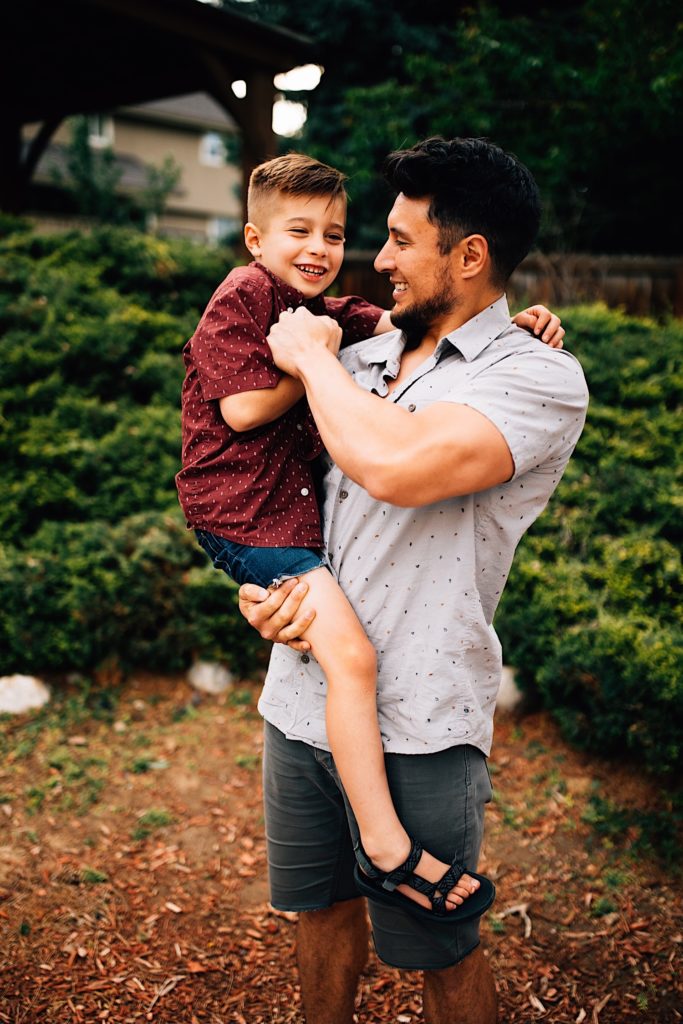 A father holds his son and smiles at him while trying to tickle him as the sun smiles at him