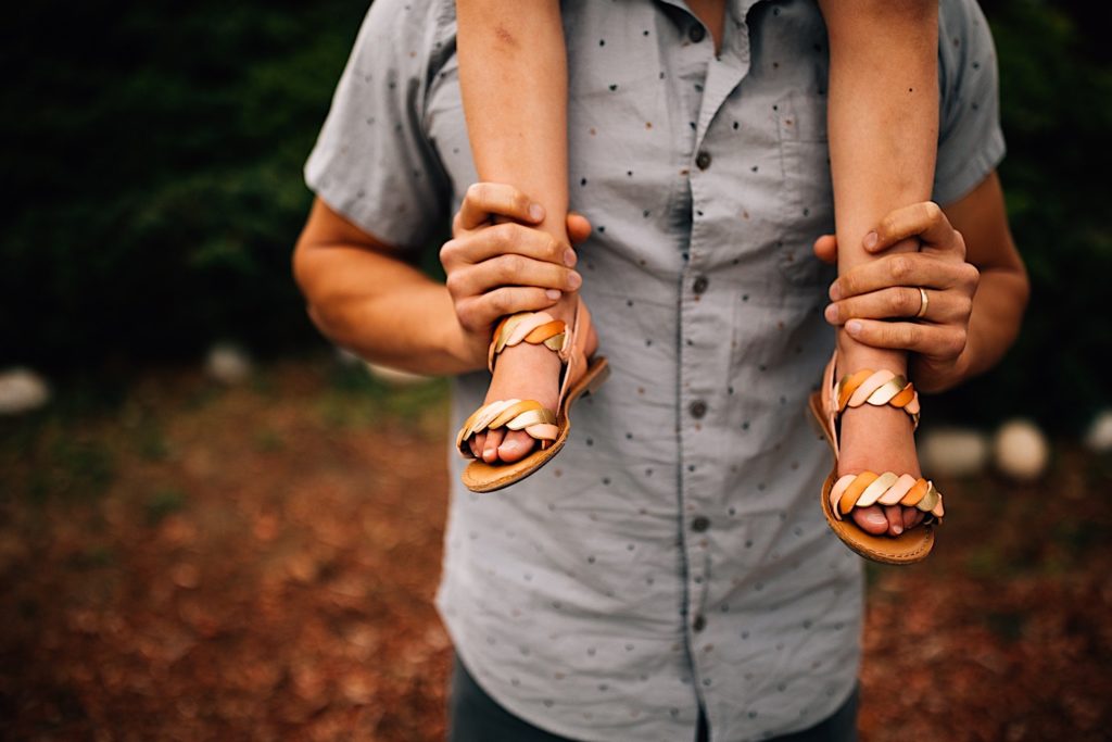 Photo of a mans torso, his daughter is sitting on his shoulders with only her legs visible as he holds her ankles.