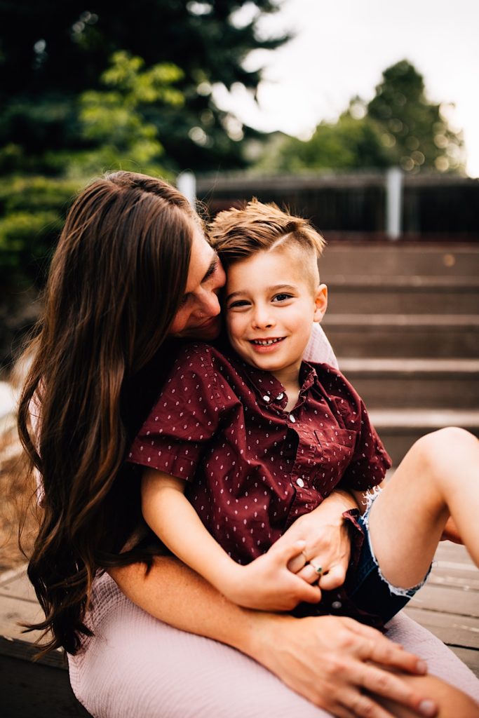 A child sits on his mothers lap outside and smiles at the camera while she holds him and smiles at him