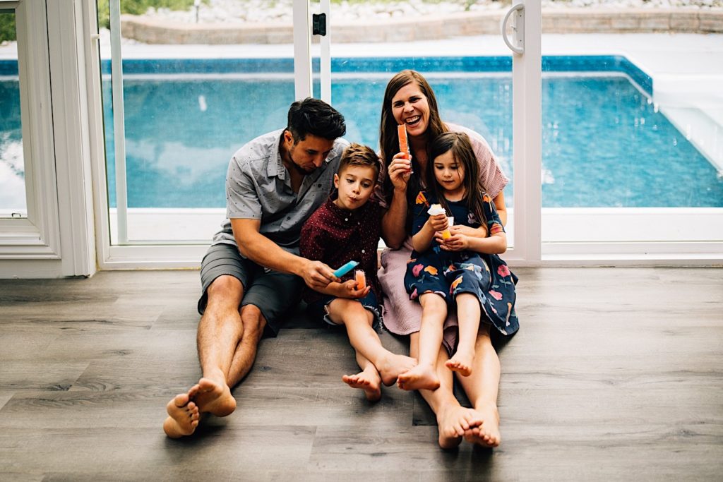 A family sits inside on the floor together eating freeze pops with their backs against a glass door with a pool behind them. The mother smiles and laughs at the camera while the others look at their freeze pops