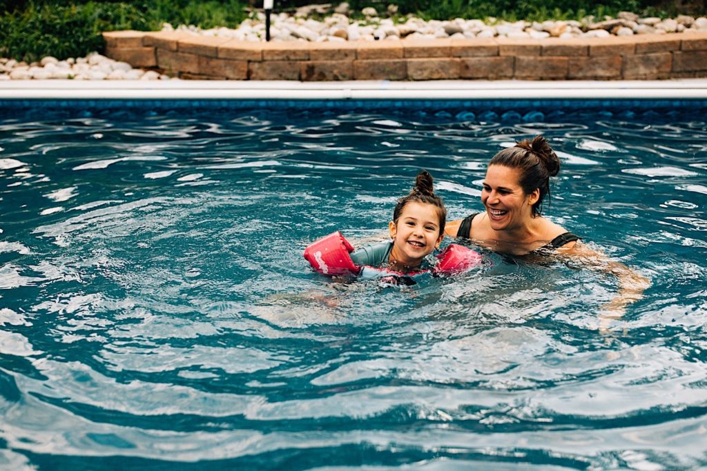 A child in a pool wearing floaties smiles at the camera as her mother swims next to her and smiles at her