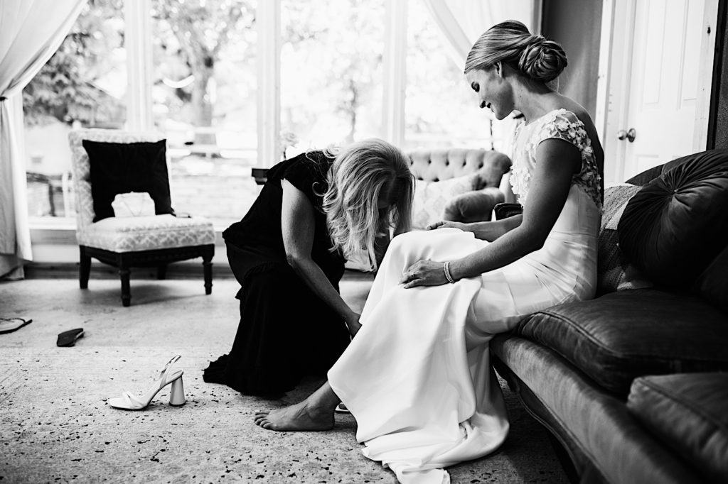 A black and white image of the brides mom helping her get on her wedding shoes in the getting ready space at River Bend in Colorado.