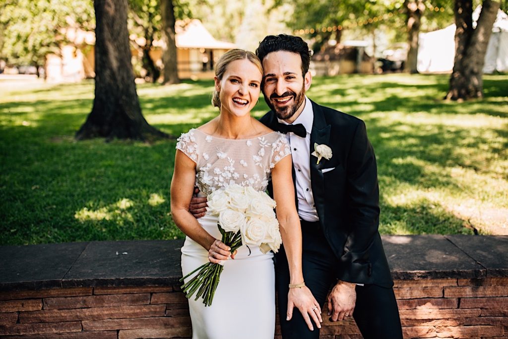 Bride and groom sit on a brick wall while smiling at the camera the morning of their wedding at River Bend in Colorado