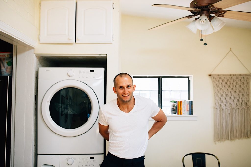A man in the laundry room of a home adjusting his shirt while smiling at the camera