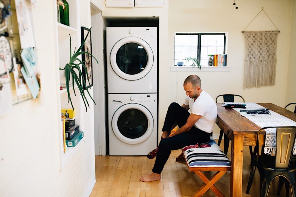 A man in a laundry room of a home sits at a table as he puts on his socks