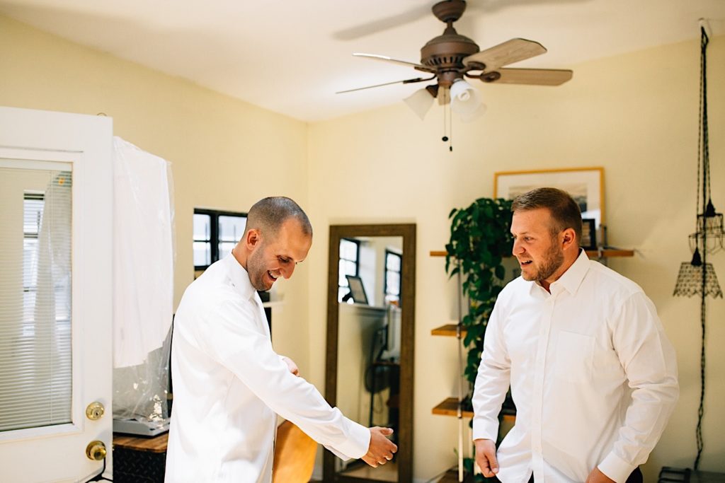 A man looks down to adjust the sleeve of his dress shirt while his friend stands next to him as they both laugh