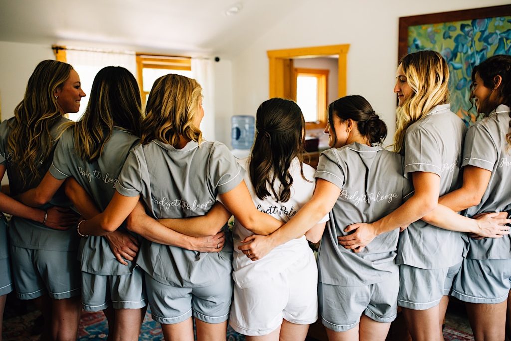 Bride and bridesmaids embrace and face away from the camera wearing matching outfits