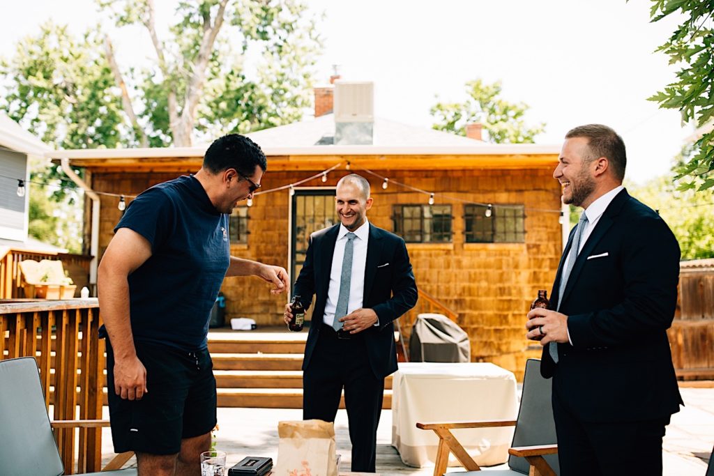A groom stands outdoors with his friends as they smile and laugh together, the groom and best man are dressed in suits and holding beer while the third man is wearing casual clothes