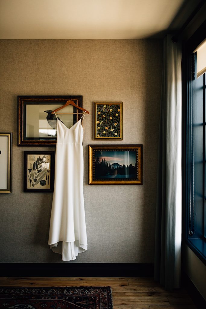 A white wedding dress hangs on a wall with framed pictures behind and a window next to it