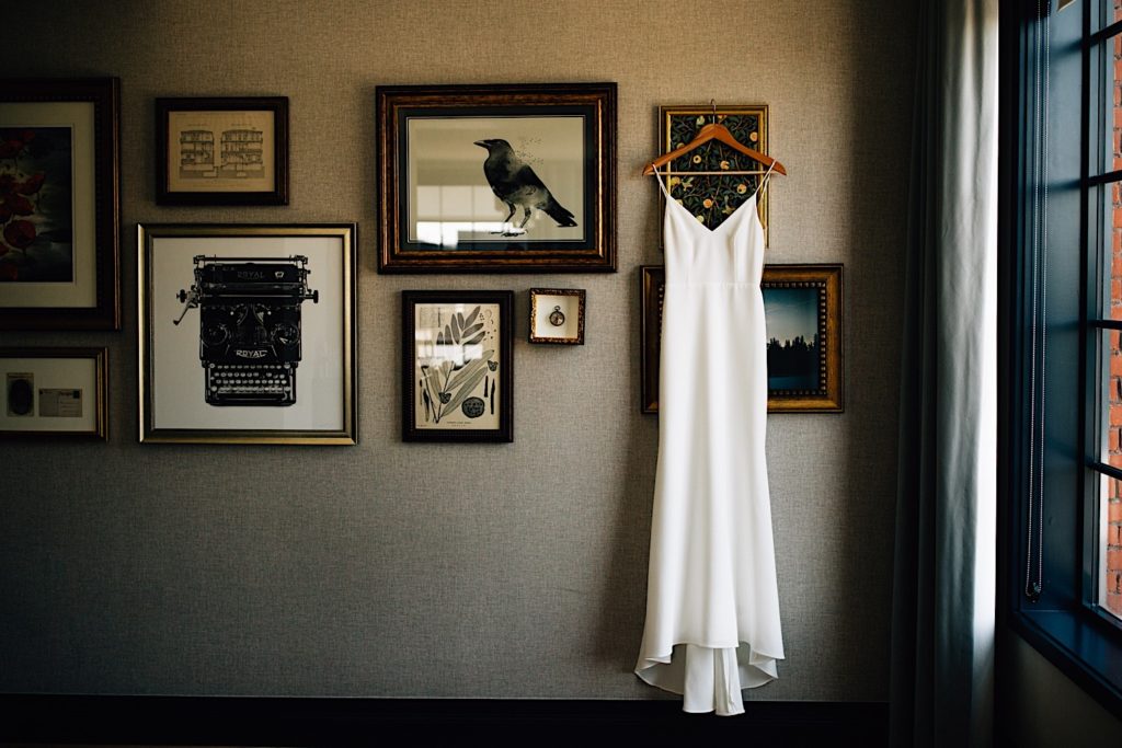 A white wedding dress hangs on a wall with framed pictures behind and a window next to it