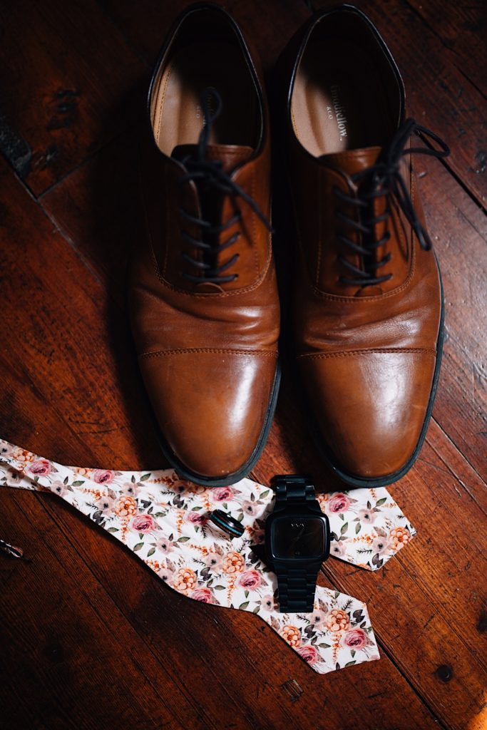 Top down photo of a wedding ring, watch, flower bowtie and brown dress shoes