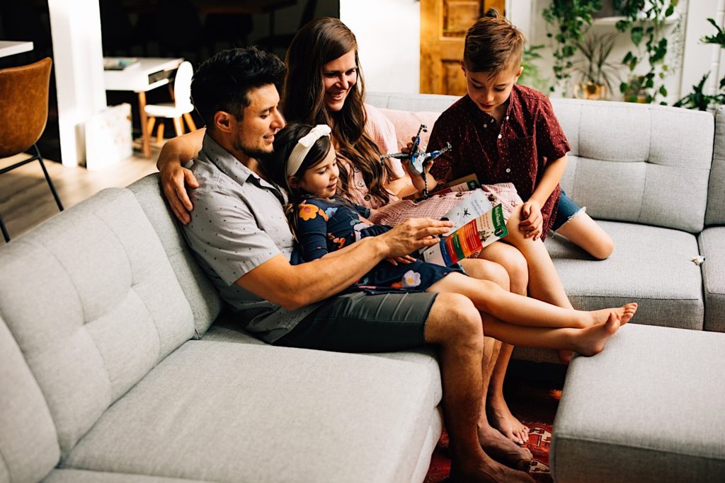 A father and mother sit on a couch in their home with their son and daughter reading a book and playing with Legos.