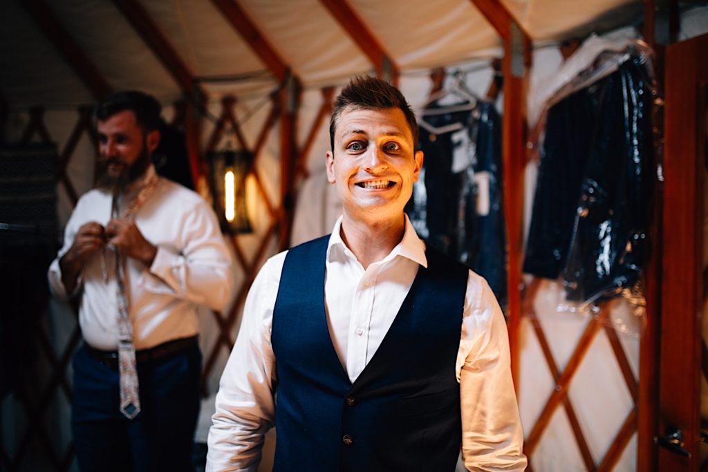 Groom smiles at the camera while a groomsmen adjusts his tie in the background 