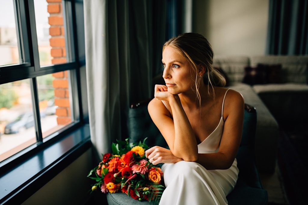 Woman wearing a wedding dress sits on a chair with her bouquet next to her and looks out of a window while resting her head on her hand, 