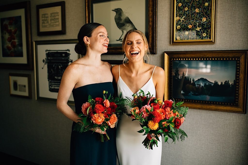 Bride and bridesmaid stand next to each other wearing their dresses, they're each holding bouquets and laughing with one another in a room with framed pictures behind them