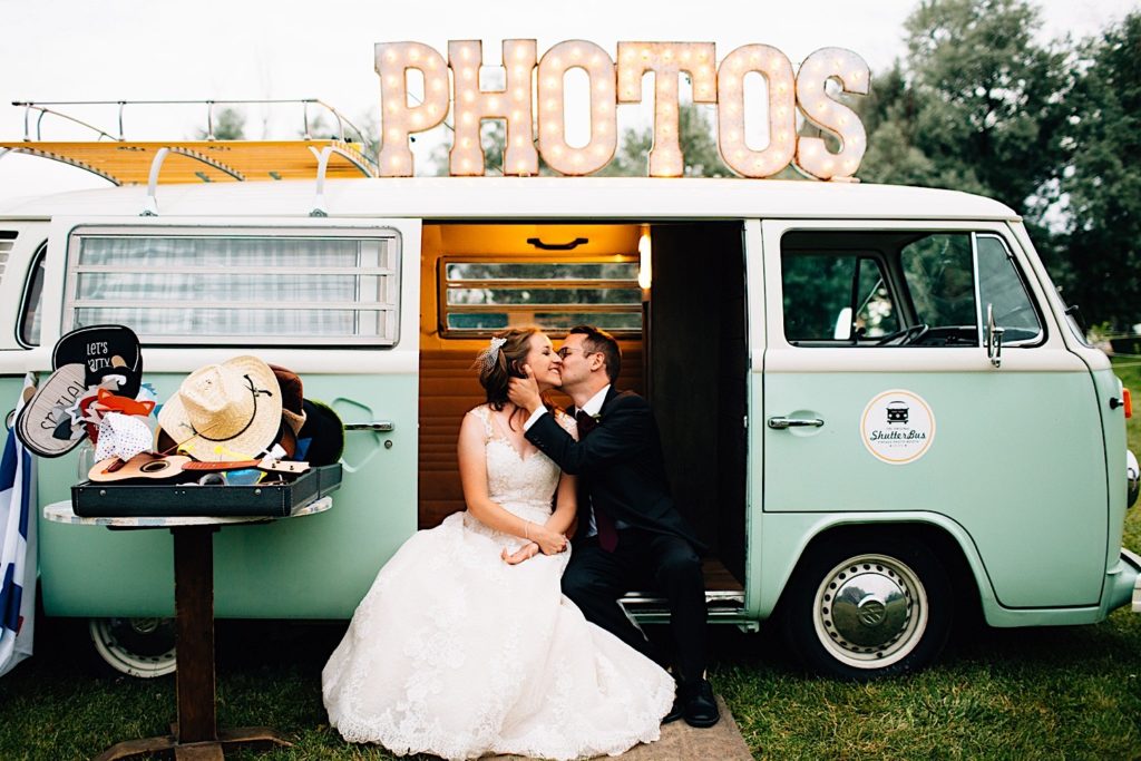 A bride and groom are seated in the side door of a classic VW van with a giant sign atop the van that reads "PHOTOS"