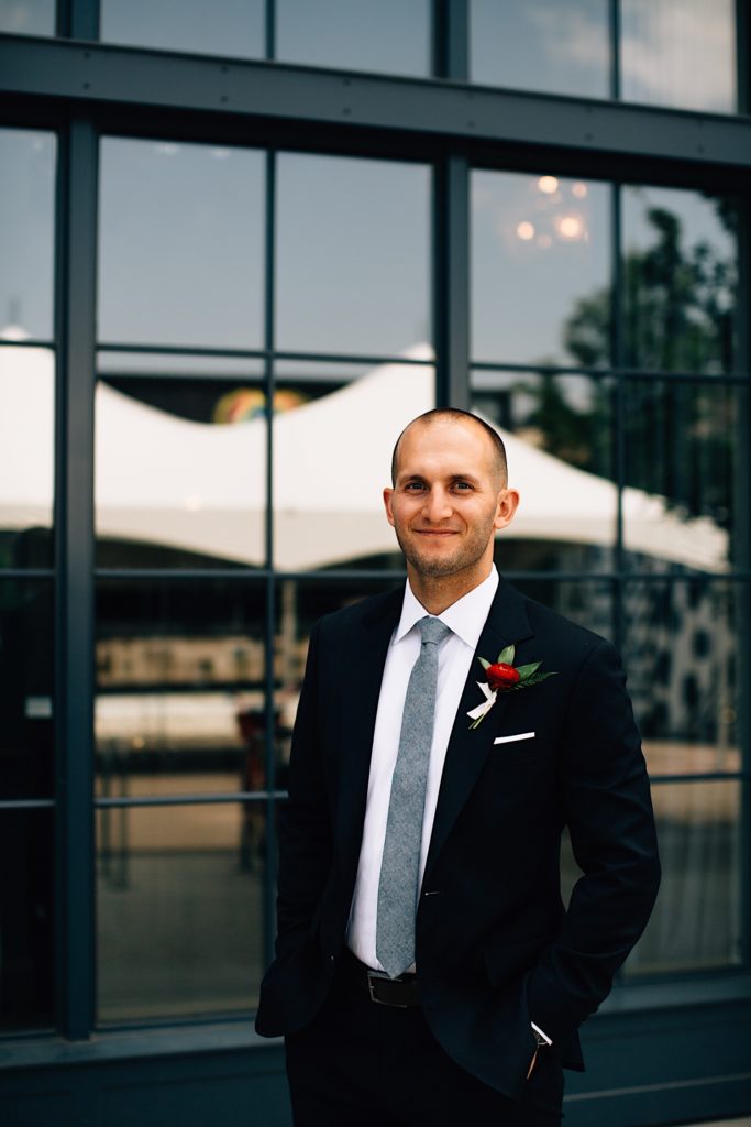 Man wearing a suit and tie with a red flower smiles at the camera in front of a building with lots of windows before his wedding