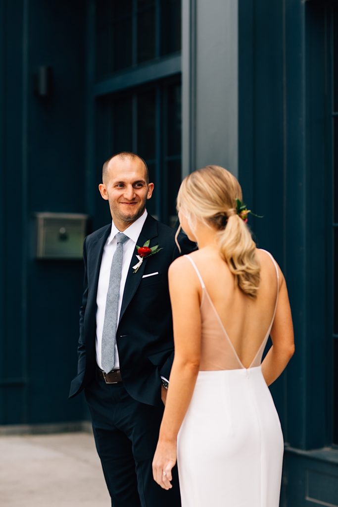 A groom smiles at his bride to be as she walks towards him, they're dressed in their wedding suit and dress respectively