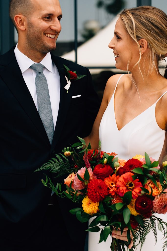A bride and groom stand next to each other and smile at one another while wearing their wedding attire, the bride is also holding a flower bouquet and they're standing in front of windows