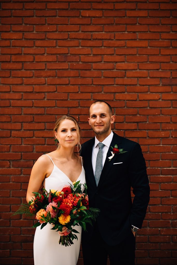 A bride and groom stand next to each other in front of a brick wall while wearing their wedding attire as they smile at the camera