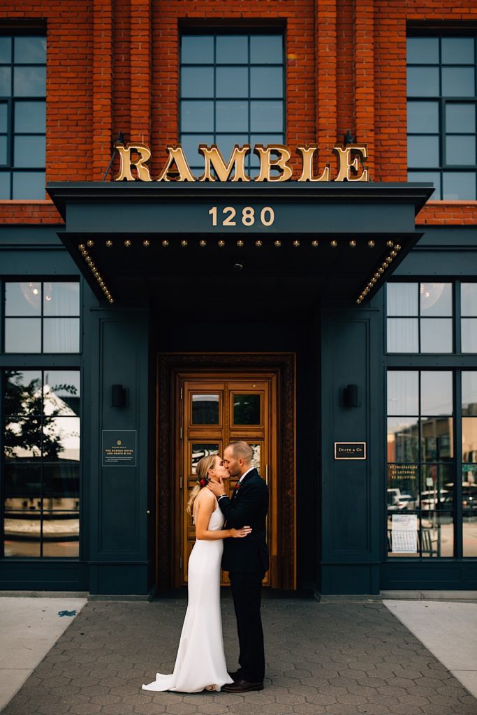 A bride and groom kiss in front of a building while wearing their wedding attire. The building reads "Ramble 1280"