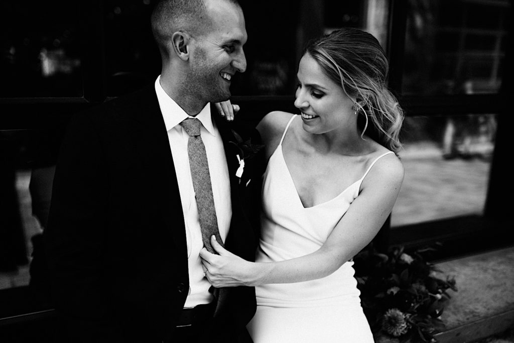 Black and white photo of a bride and groom standing next to each other wearing their wedding attire, they smile as the bride adjusts the grooms tie