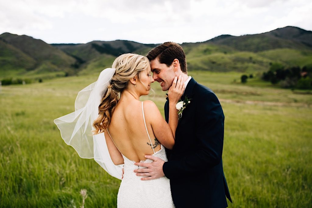 A bride and groom stand facing one another wearing their wedding attire with rolling fields and mountains behind them. 
