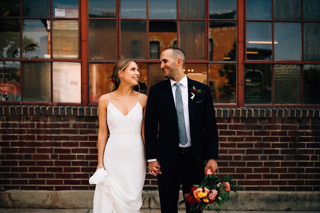 Bride and groom stand next to each other and hold hands while looking at one another wearing their wedding attire, they're standing in front of a glass and brick building and the groom is holding a flower bouquet