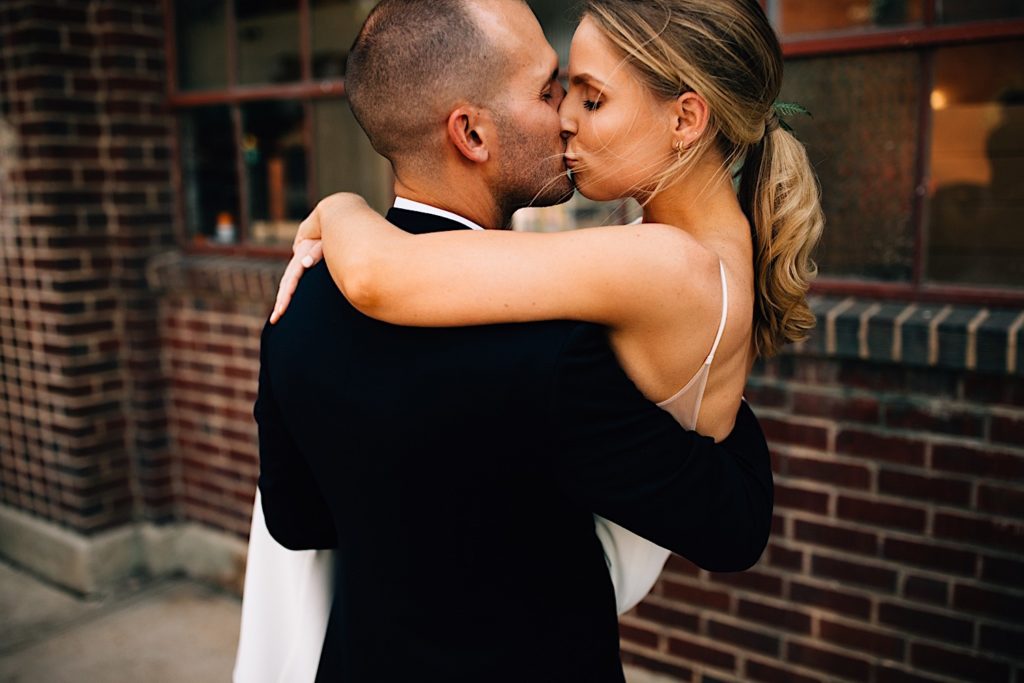 A groom carries his bride with his back facing the camera, they both kiss as their wearing their wedding attire outside of a glass and brick building