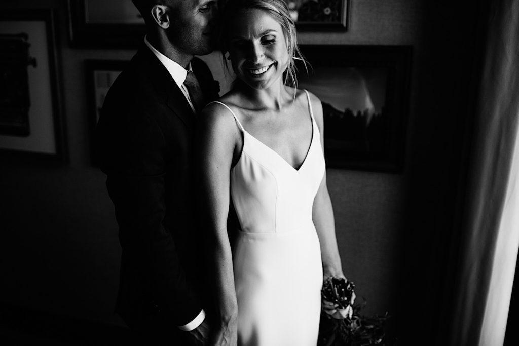 Black and white photo of a bride and groom in a room with framed photos on the wall, the groom stands behind the bride while they both smile
