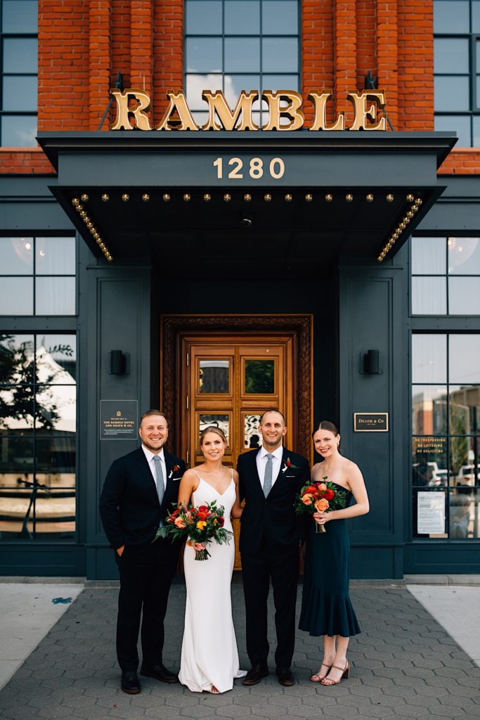 A bride, groom, best man and maid of honor all stand together wearing their wedding attire outside a building with "Ramble 1280" on a sign above them