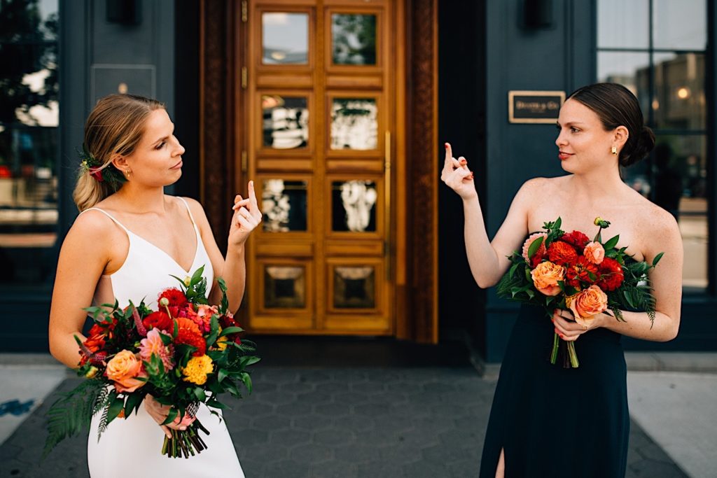 Bride and her maid of honor look at one another and jokingly flip each other off while each holding a flower bouquet outside the wedding venue