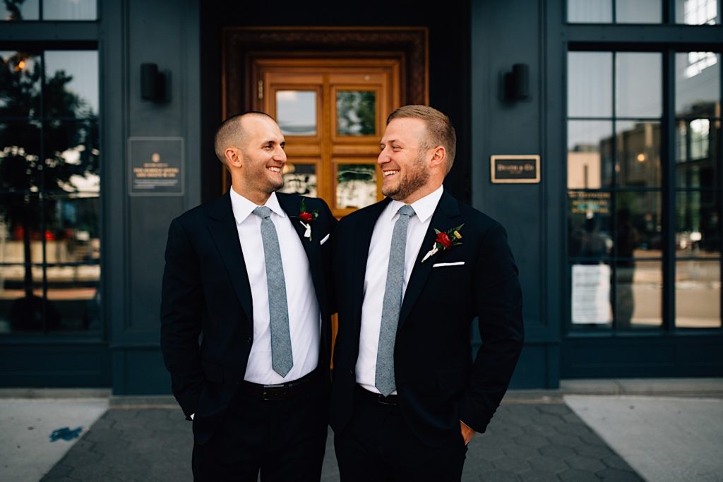 Groom and best man stand next to one another and smile as they look at one another outside the wedding venue