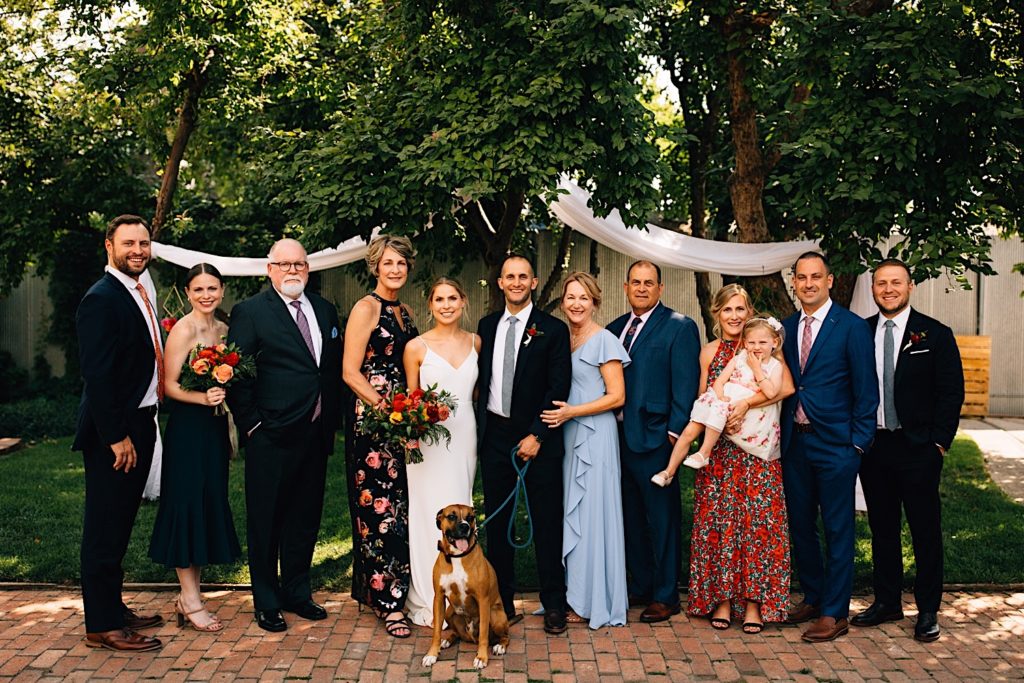 Photo of a bride and groom with their families all lined up next to them and their dog sitting in front of them, they're outdoors and have trees with white cloth behind them