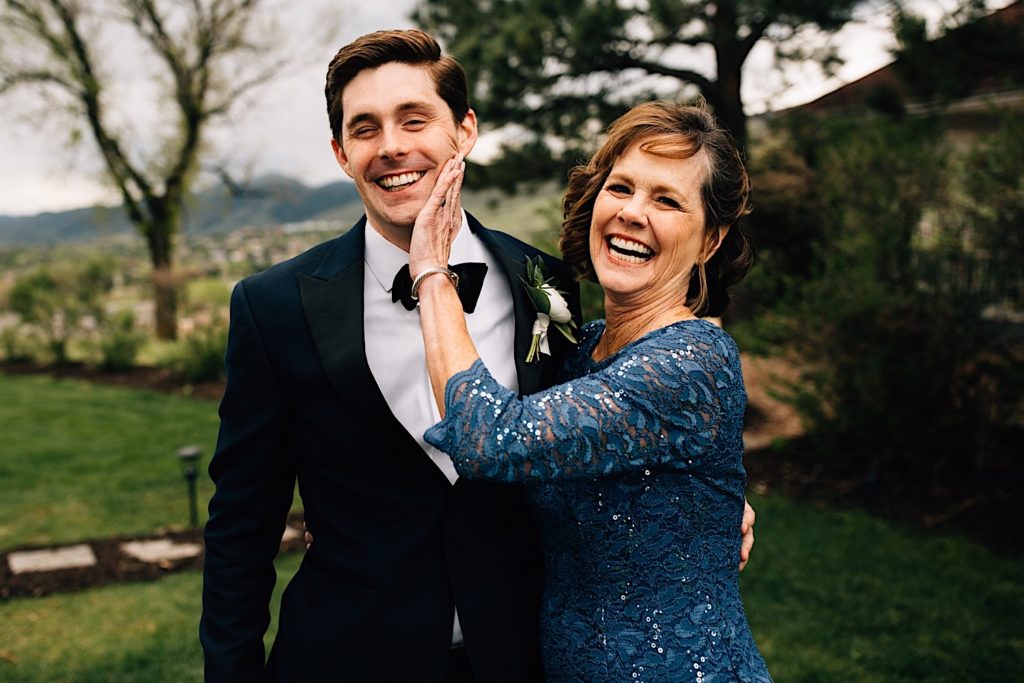 A groom and his mother stand side by side and smile at the camera as the mother puts her hand on her sons cheek.