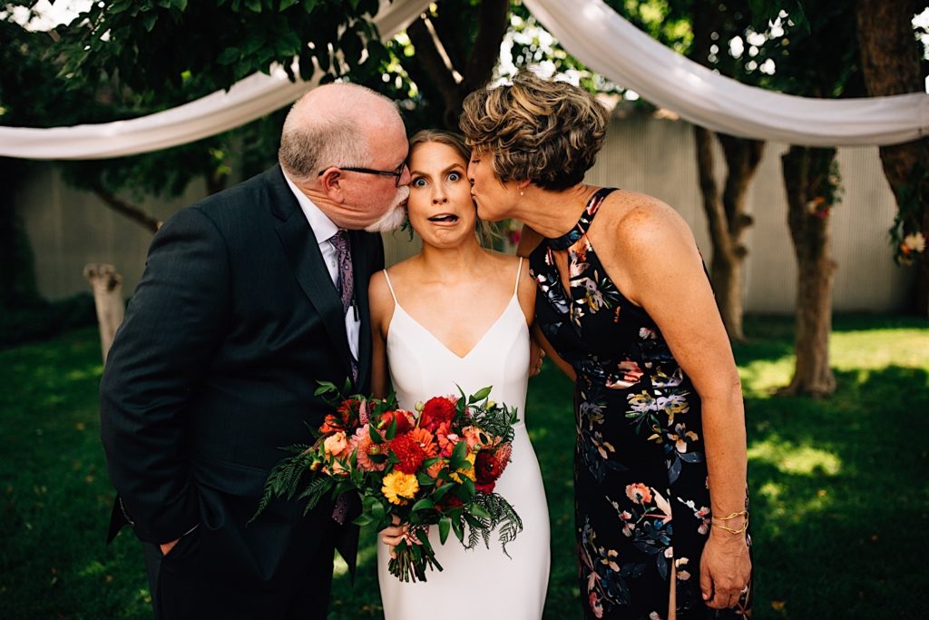 A bride stands in between her parents, her parents both kiss her on opposite cheeks as she makes a funny face, they're dressed for the wedding and standing outside at the venue