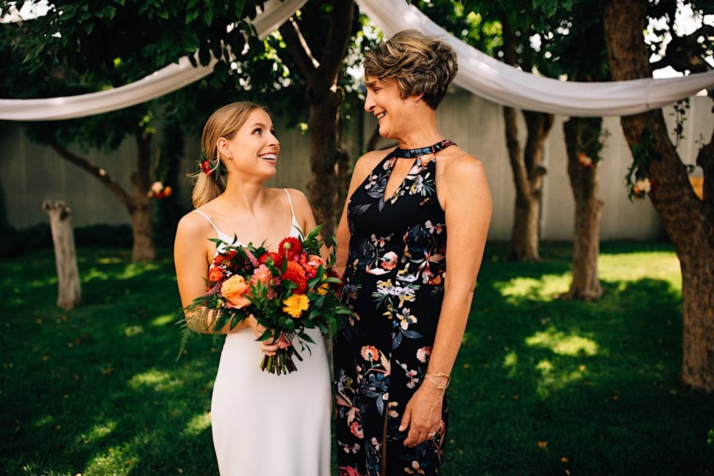 Bride and her mother stand next to one another and look at each other smiling, they're outdoors at the wedding venue