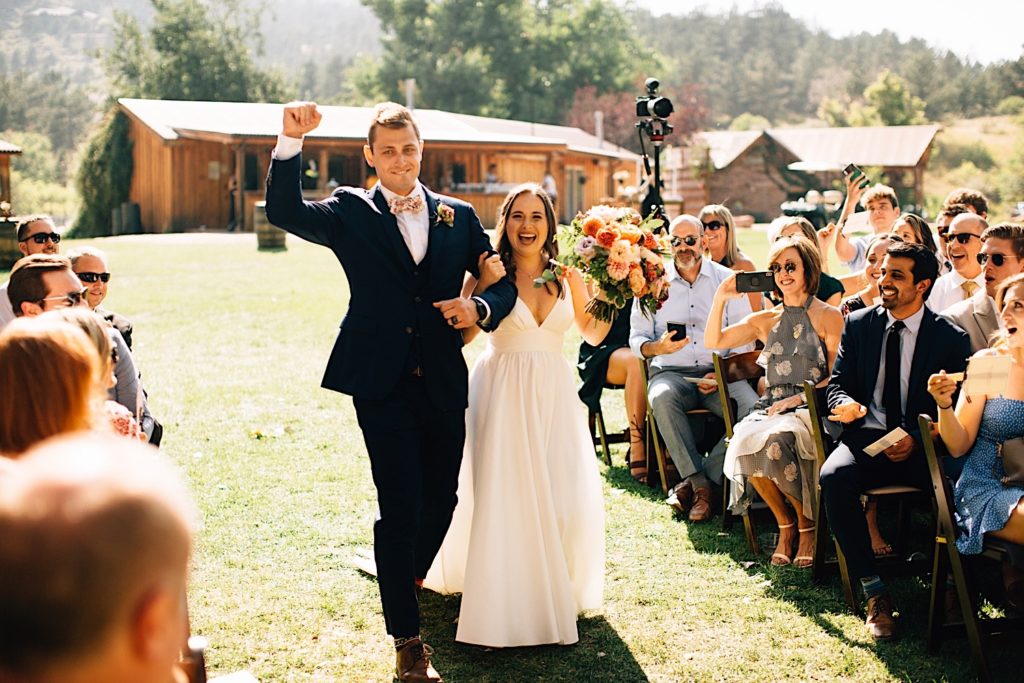 Bride and groom walk down the aisle together at their Planet Bluegrass wedding with their guests smiling at them, the bride is holding a bouquet while the groom has a fist in the air