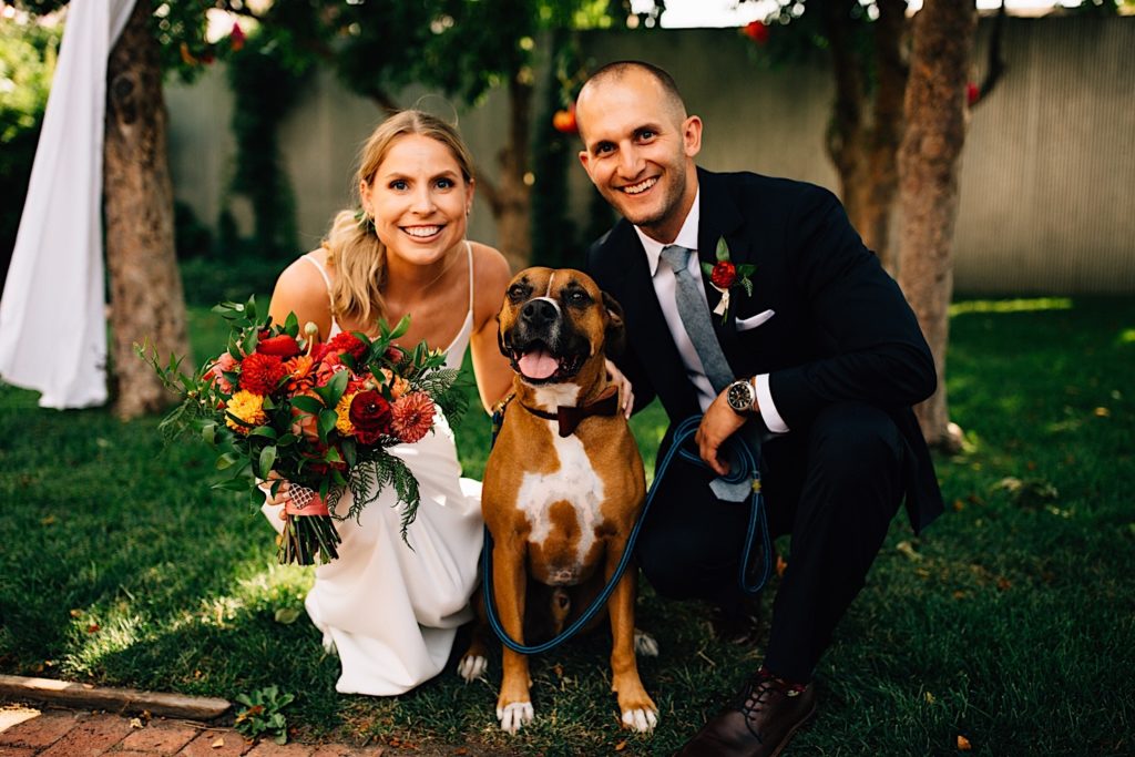 A bride and groom both crouch down and smile at the camera while their dog sits in between them looking at the camera. The bride and groom are dressed for their wedding, the bride is holding a flower bouquet and they are outdoors at their wedding venue