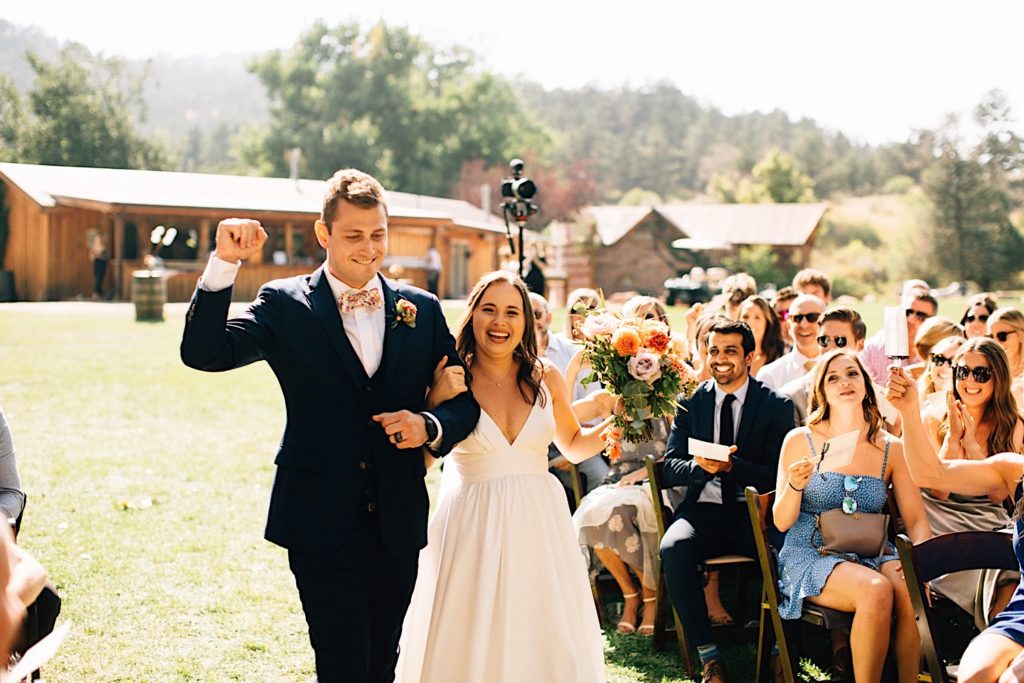 Bride and groom walk down the aisle together at their Planet Bluegrass wedding with their guests smiling at them, the bride is holding a bouquet while the groom has a fist in the air