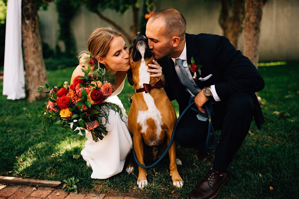 A bride and groom both crouch down kiss their dog on opposite cheeks while their dog sits in between them looking at the sky. The bride and groom are dressed for their wedding, the bride is holding a flower bouquet and they are outdoors at their wedding venue