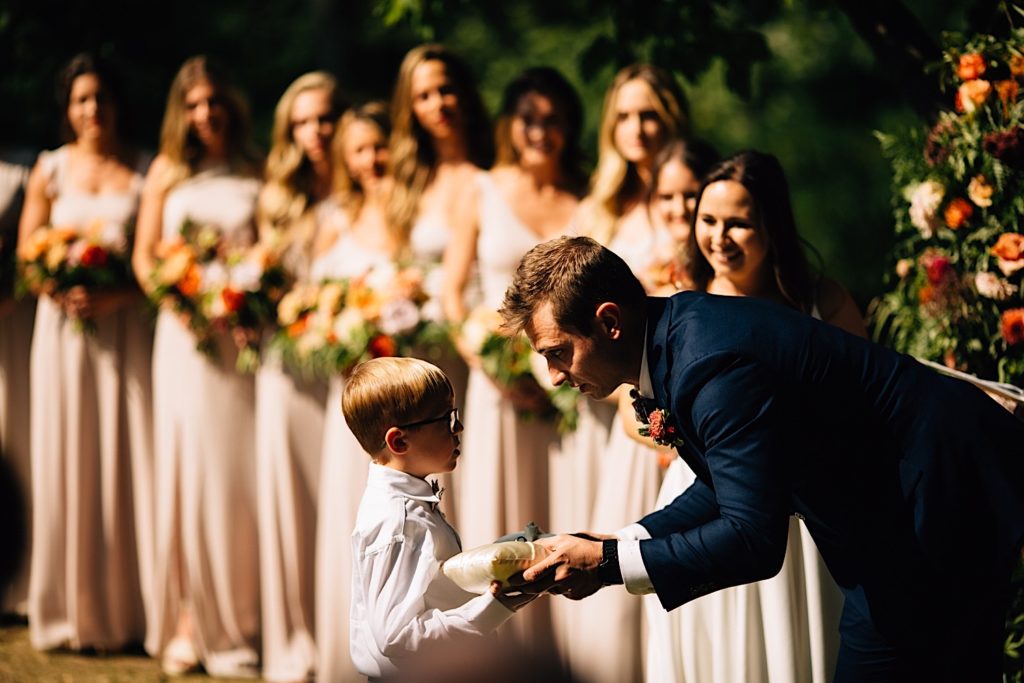 Groom accepts the ring from the ring bearer with the bride and her bridesmaids in the background