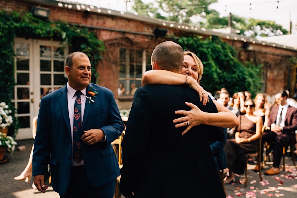 The groom at a wedding has his back turned to the camera as his mother hugs him while his father stands next to them and smiles, guests of the wedding are seated in the background