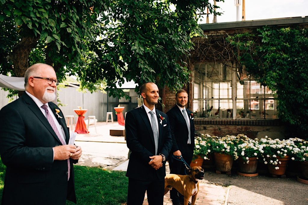 A groom stands and smiles with his hands clasped while his best man and dogs stand on his left and the marriage officiant stands on his right, they are outdoors at their intimate wedding venue