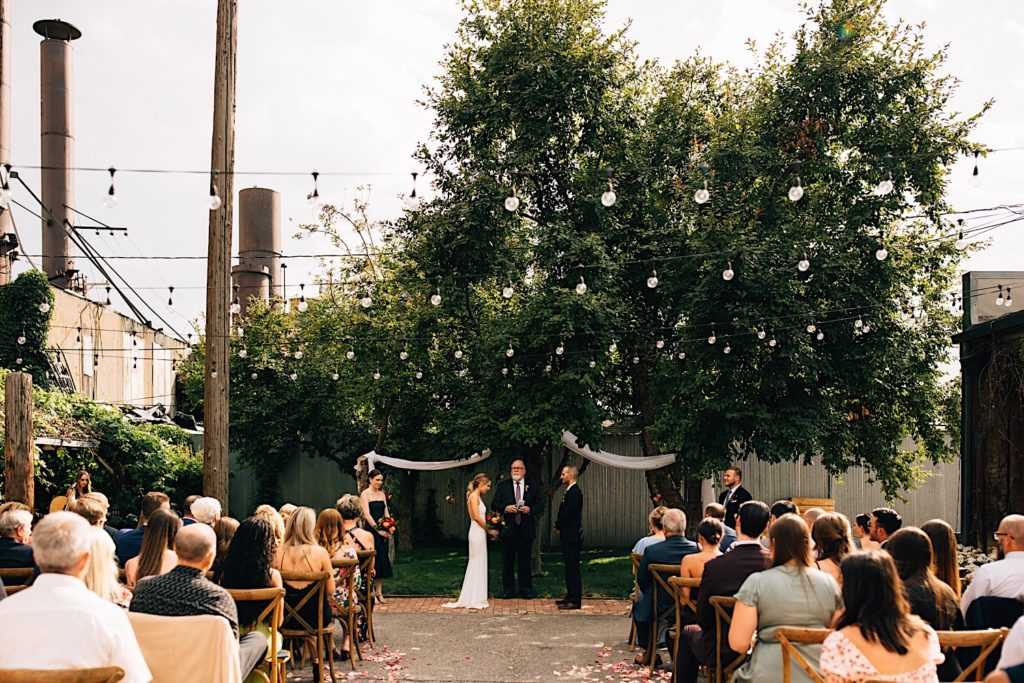 An intimate wedding ceremony, the bride and groom stand with their officiant while their guests are seated and the best man and maid of honor stand on either side of the couple. The intimate wedding venue is outdoors and has string lights above the guests and trees behind the couple