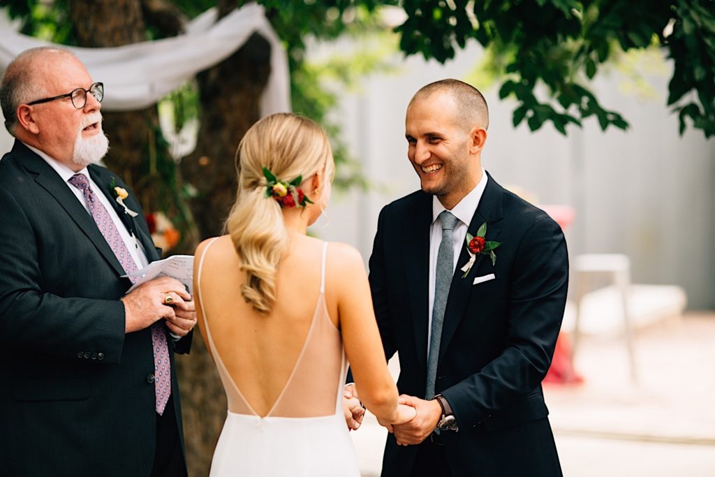 A groom smiles as he holds his brides hands and looks at her, the bride has her back turned to the camera, the officiant stands to the left of the bride and speaks to them