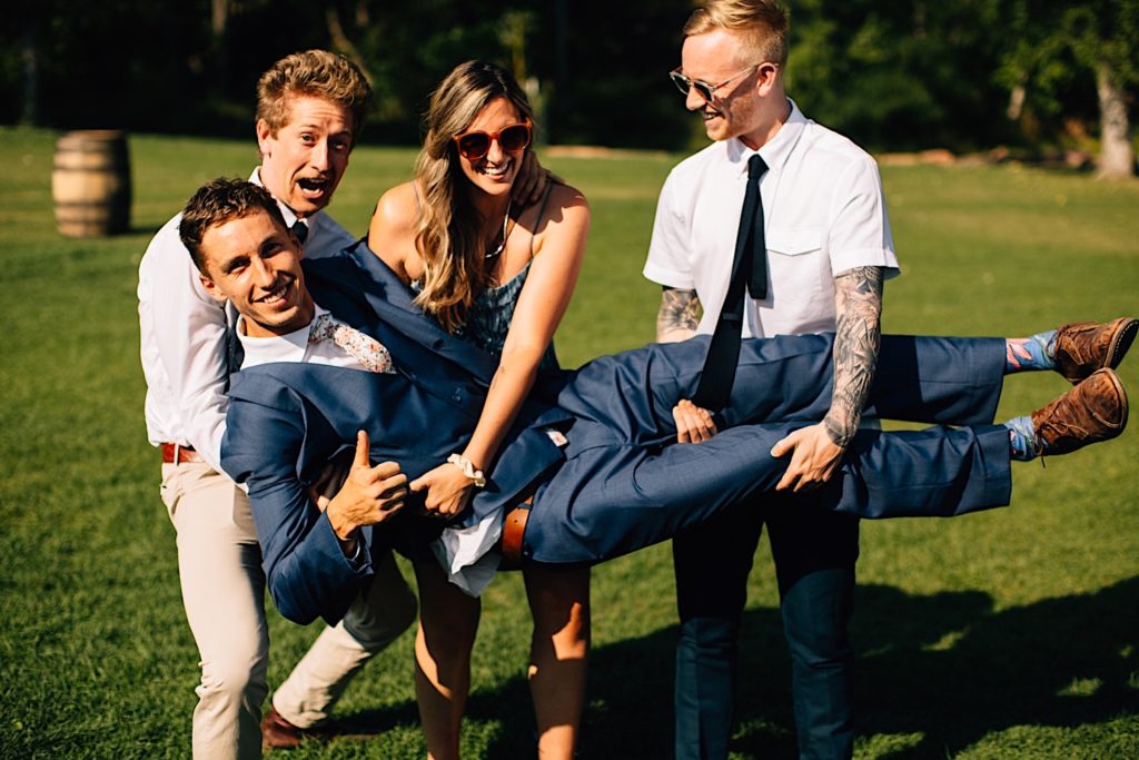 Groomsmen gives a thumbs up as his friends hold him up and smile
