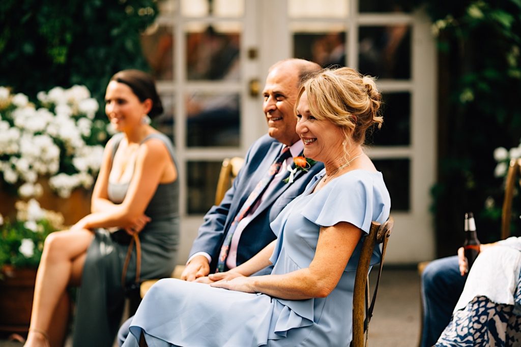 Parents seated at an outdoor intimate wedding with guests around them, there are white double door and some flowers in the background